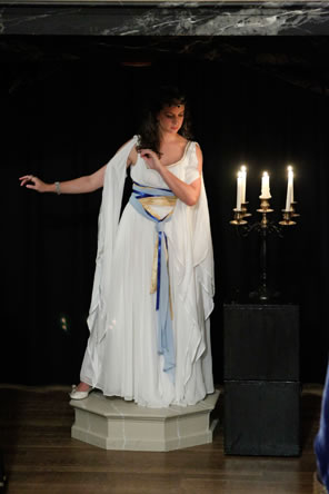 Hermione in Gree-like gown posing as the statue, on a pedestal with a candelabra next to her