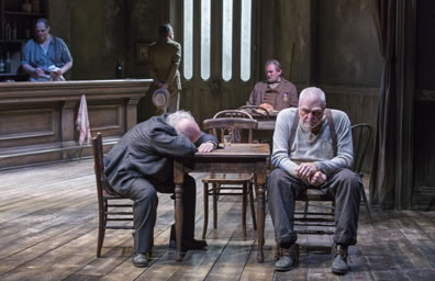 At center, Slade sitting at a right side of table but facing forwrd, left hand gripped over right wrist, slumped forward, staring straight ahead, work pants with suspenders and long-johns shirt, across from him Hugo in gray suit has his head on his folded arms asleep on the table top, in the background at another table Piet sits in three-piece brown british outfit with a medal on his left pocket, Captain Lewis stands, holding hat in his hands behind his back looking out the door, and to the left Rocky the bartender in blue shirt cleans glasses.