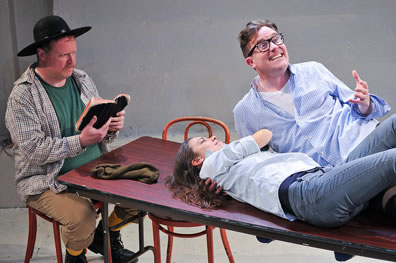 Olivia in blue shirt and jeans lies on table with Sebastian, raised upon one elbow and expressing himself with his left hand. At the head of the table sits the priest in checkered shirt ove green t-shirt and wearing brown pants and yellow socks, a black padre's hat and holding upen a tattered Bible