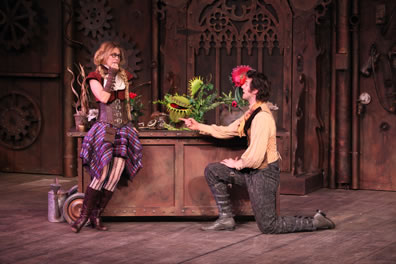 Luciana in red hair with pig tails leans against a copper wheeled table covered with exotic plants. She's wearing red bodice, leather corset, leather wrist bands, purple plaid skirt fallen away to reveal exotically stocking legs and brown boots. Antipholus is across from her, kneeling on one knee in gray pants and boots, a yellow shirt and gold vest. In the background is the wood door amid the gadgetry of the back wall. 