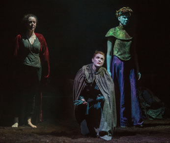 Henry IV in silver tunic, black pants, bare feet, and red robe, Richard wearing the crown, in white face, gold brocade blouse and purple dress, Henry V kneeling, gloves in hand, gray cloak over her.