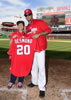 Desmond in red W basball cap, sunglasses, windbreaker and white uniform hat next to big-smiling Sarah in baseball cap, red shirt, blue jeans, and spider sneakers, both holding the red baseball jersey with "DESMOND 20" on the back. This is at home plate with the baseball field, scoreboard, and Red Porch cafe and beautiful blue skies with whispy clouds in the background.