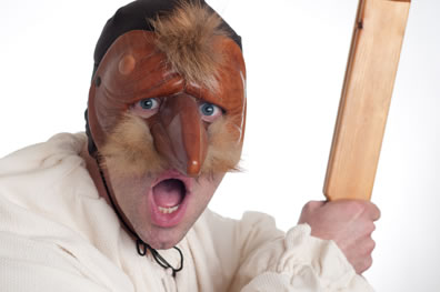 Wilson in brown mask with long sharp nose and tuffs of brown hair as a mustache and erupting from the top of the mask, his mouth open, his blue eyes staring through the eye slites, black cap tied around his neck, his white smock with frilly cuffs, and holding to his side the wood clapper in his hand. 