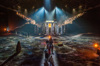 A line of people walk down a central stone path through a heath toward the Stonehenge-end of Macbeth's play space, with spotlights spraying out from the ceiling onto the two grandstands of people