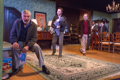 Leontes in casual sport jacket, open collar shirt, nice pants sitting on toy box, Lincoln Log set at his feet, Persian rug on the floor, closet, bar cabinet, poster, and rest of dining room in background, Lord carrying the swaddling clothes around a baby, Antigonis walking behind him, and two other lords over by the dining room table