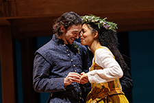 William in a blue Elizabethan patterned vest jacket and shirt snuggles and holds hands with Agnes, wearing a white shirt and brown patterned dress and a crown of flowers.