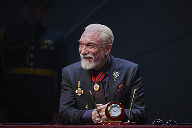 Patrick Page as King Lear, wearing a dark blue suit with purple tie, his jacket adorend with medals and a med on a red and blue ribbon around his neck, sits at a desk, arms folded and sporting a gentle smile. In front of him on the desk is a clock-centered pen stand. Photo by DJ Corey Photography.