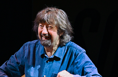 Sir Trevir Nunn Portrait of Sir trevor Nunn, smiling, wearinga blue denim shirt against a black background