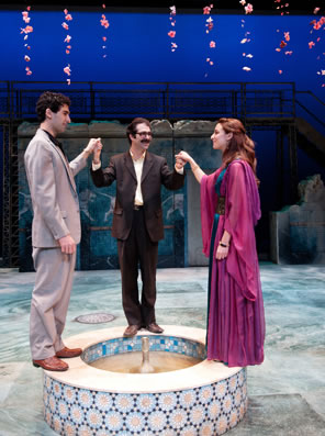 Pandarus holds the hands of Troilus in gray suit and Cressida in long red gown as they stand on a dry fountain under hanging flowers.